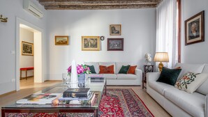 A living room with a white sofa, a coffee table with books and a vase of flowers, a red patterned rug, and a wooden ceiling. - Miracoli Apartment with View on the Canal R&R (Venecia)