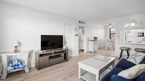 A modern living room with a flat-screen TV, white furniture, and a kitchen area in the background.