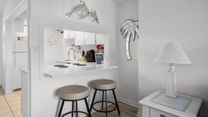 A modern kitchen with a white countertop, two stools, a decorative dolphin wall art, a palm leaf wall art, and a small table with a lamp.