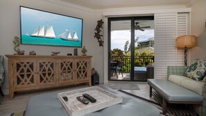 A living room with a large TV, wooden cabinet, sofa, and a balcony with outdoor furniture.