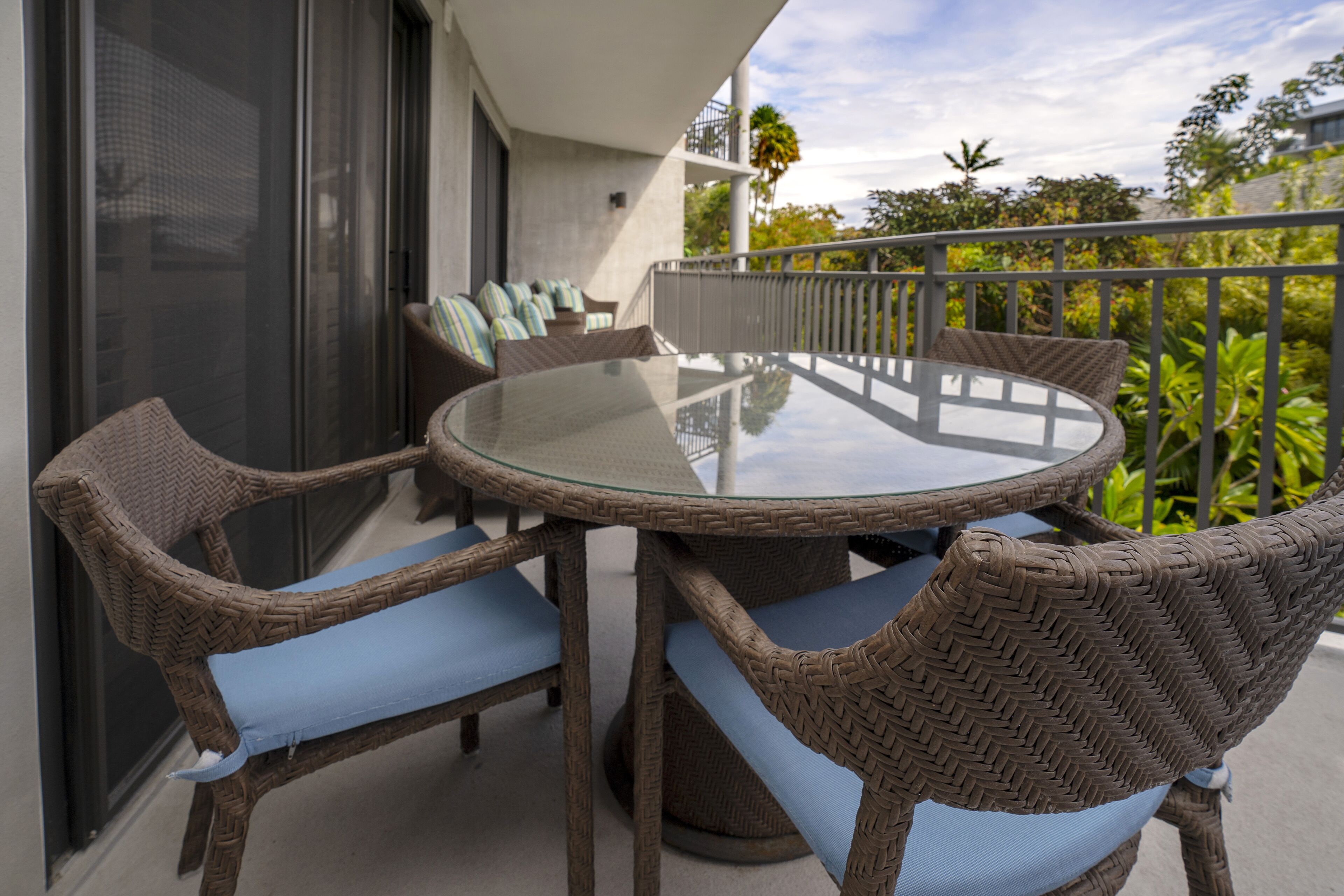 A round glass table with wicker chairs and cushions on a balcony.