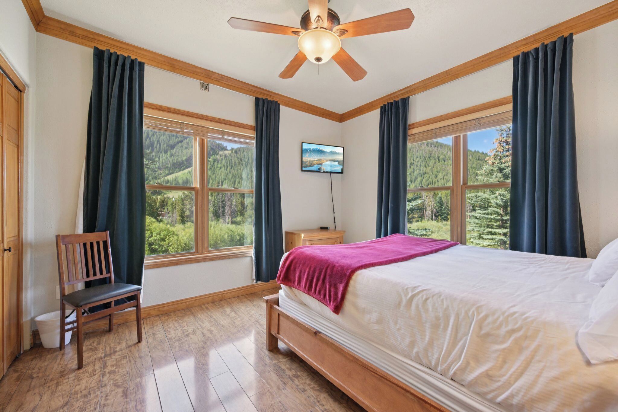 A bedroom with a bed, a wooden chair, a nightstand, a TV, and large windows with curtains.