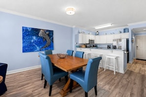 A dining area with a wooden table and blue chairs, a kitchen with white cabinets, and a blue seahorse painting on the wall.