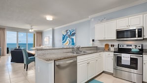 A modern kitchen with a dishwasher, microwave, and oven. There is a dining area with a table and chairs. A large window offers a view of the ocean.