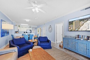 A living room with blue furniture, a wooden coffee table, a television displaying a beach view, and a clock on the wall.