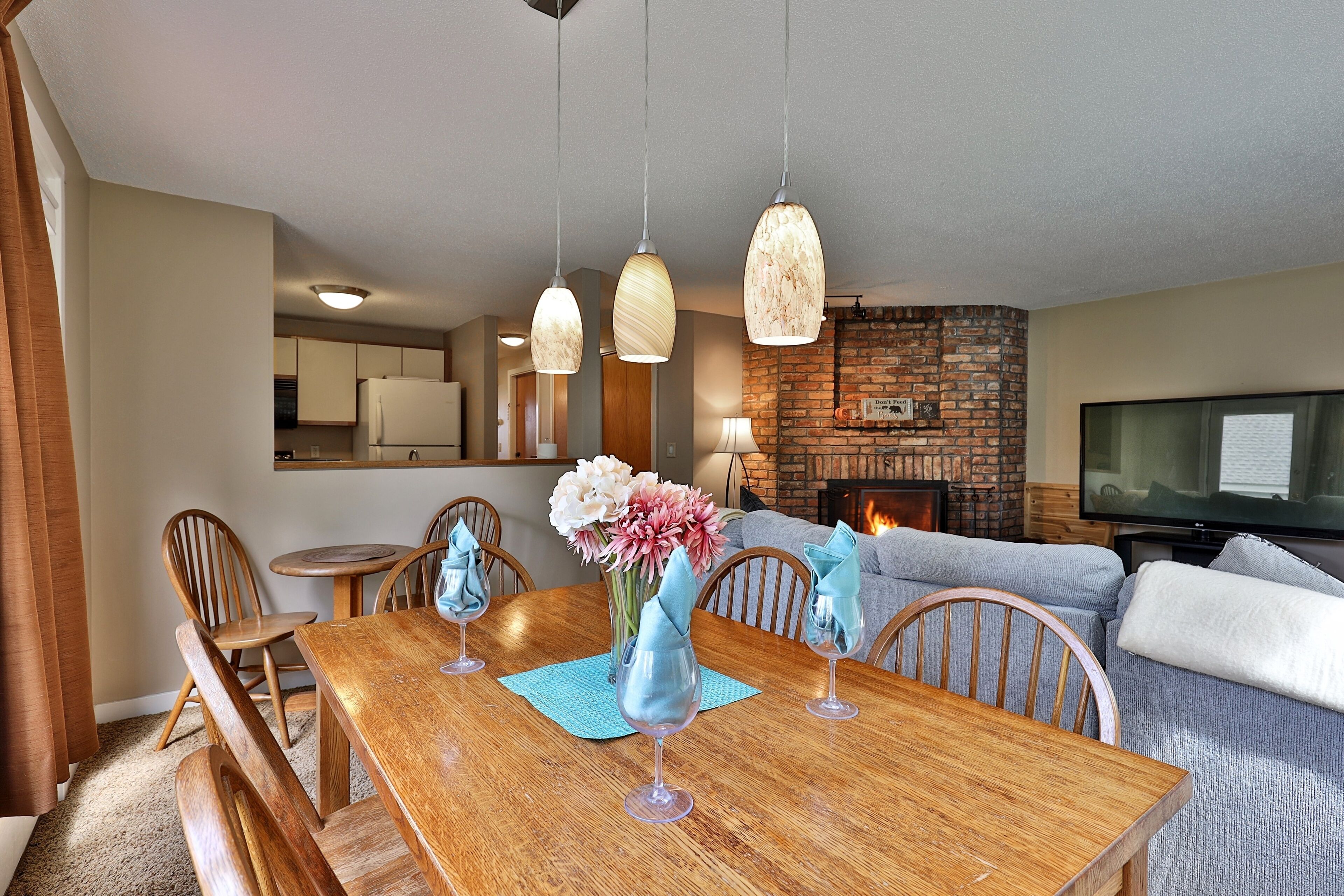 A dining area with a wooden table, chairs, and a vase of flowers. There is a brick fireplace and a television in the background.