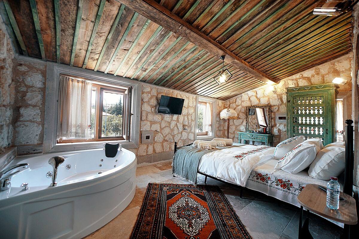 Une chambre à coucher rustique avec un mur en pierre, une grande baignoire, un lit recouvert d’une courtepointe à motifs et un plafond en bois.