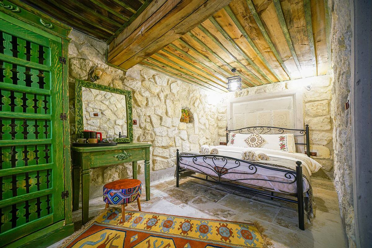 Une chambre à coucher rustique avec un mur en pierre, des poutres en bois au plafond, une porte verte, un lit avec un cadre en métal et un petit tabouret.