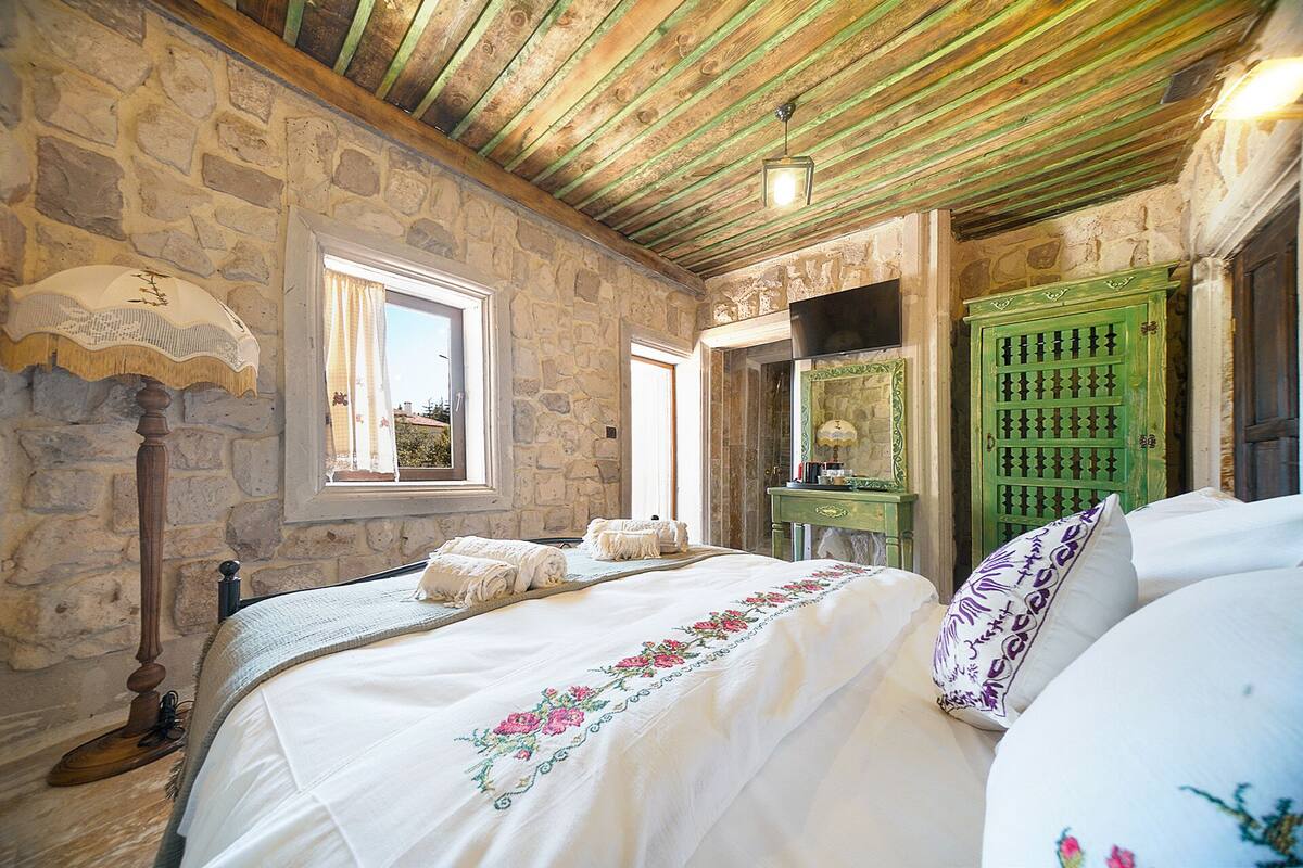 Une chambre avec des murs en pierre, un plafond en bois, un lit avec une literie à motifs floraux, une porte verte et une fenêtre avec des rideaux.