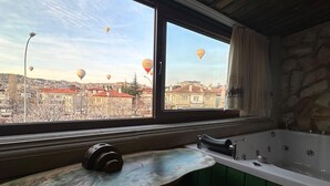 Ein Badezimmer mit einer Badewanne, einem Fenster mit Vorhängen und Blick auf eine Stadtlandschaft mit Heißluftballons.