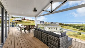 A covered outdoor area with a wooden deck, a wicker sofa, a dining table with chairs, and a hanging light fixture. - Seaglass - Karikari Peninsula Holiday Home (Kaitaia)