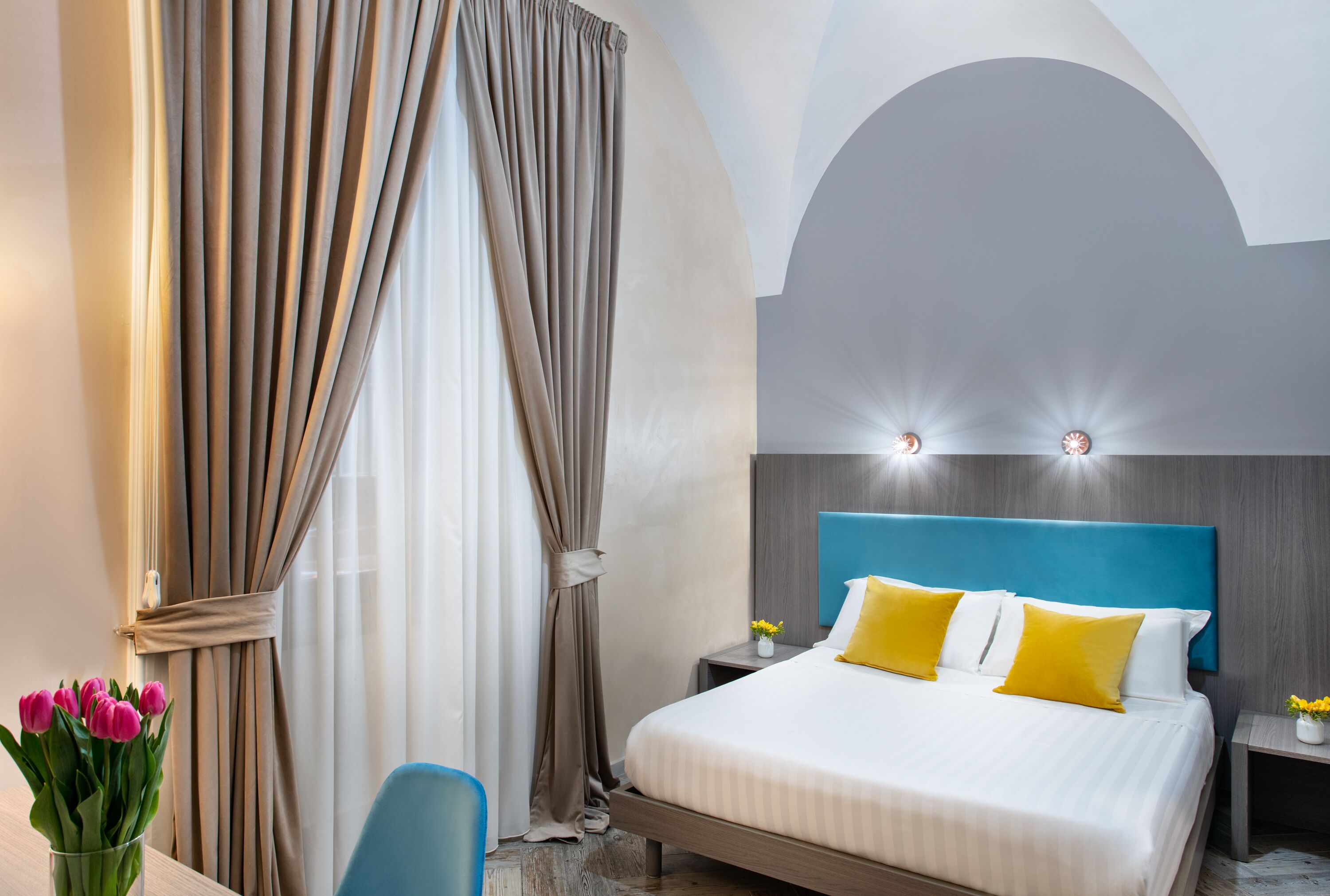A hotel room with a bed, curtains, a nightstand, and a vase of flowers.