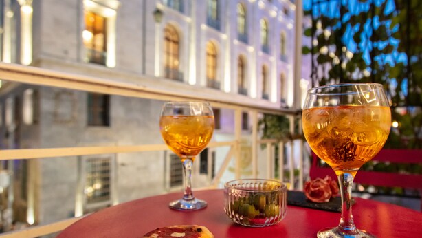 A table with two glasses of amber liquid, a small bowl of pickles, and a slice of pizza on a red table in an outdoor setting with a building in the background. - Residenza Toledo Lifestyle (Naples)