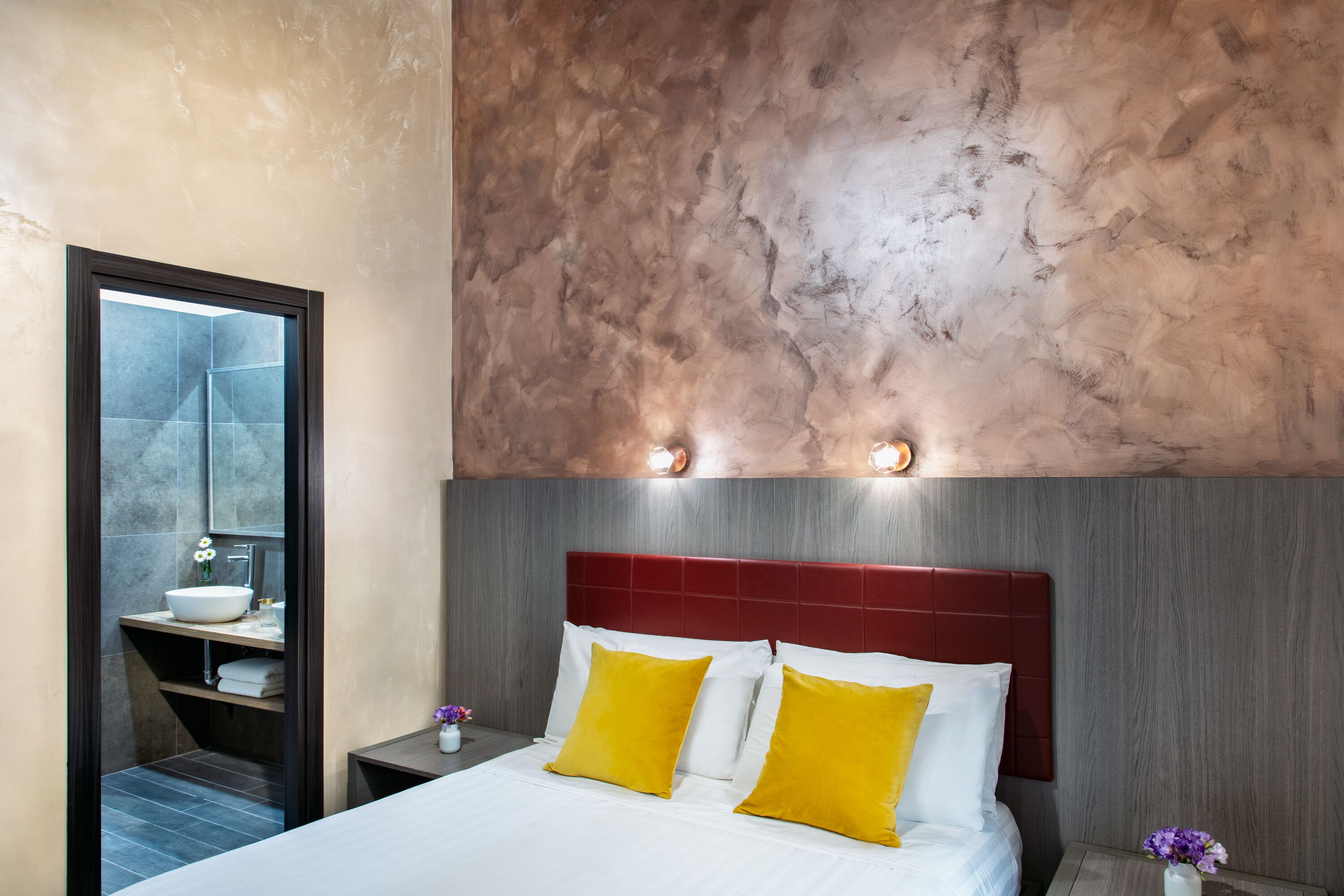 Una habitación de hotel moderna con una cama grande, cabecera roja y baño visible a través de un panel de vidrio.