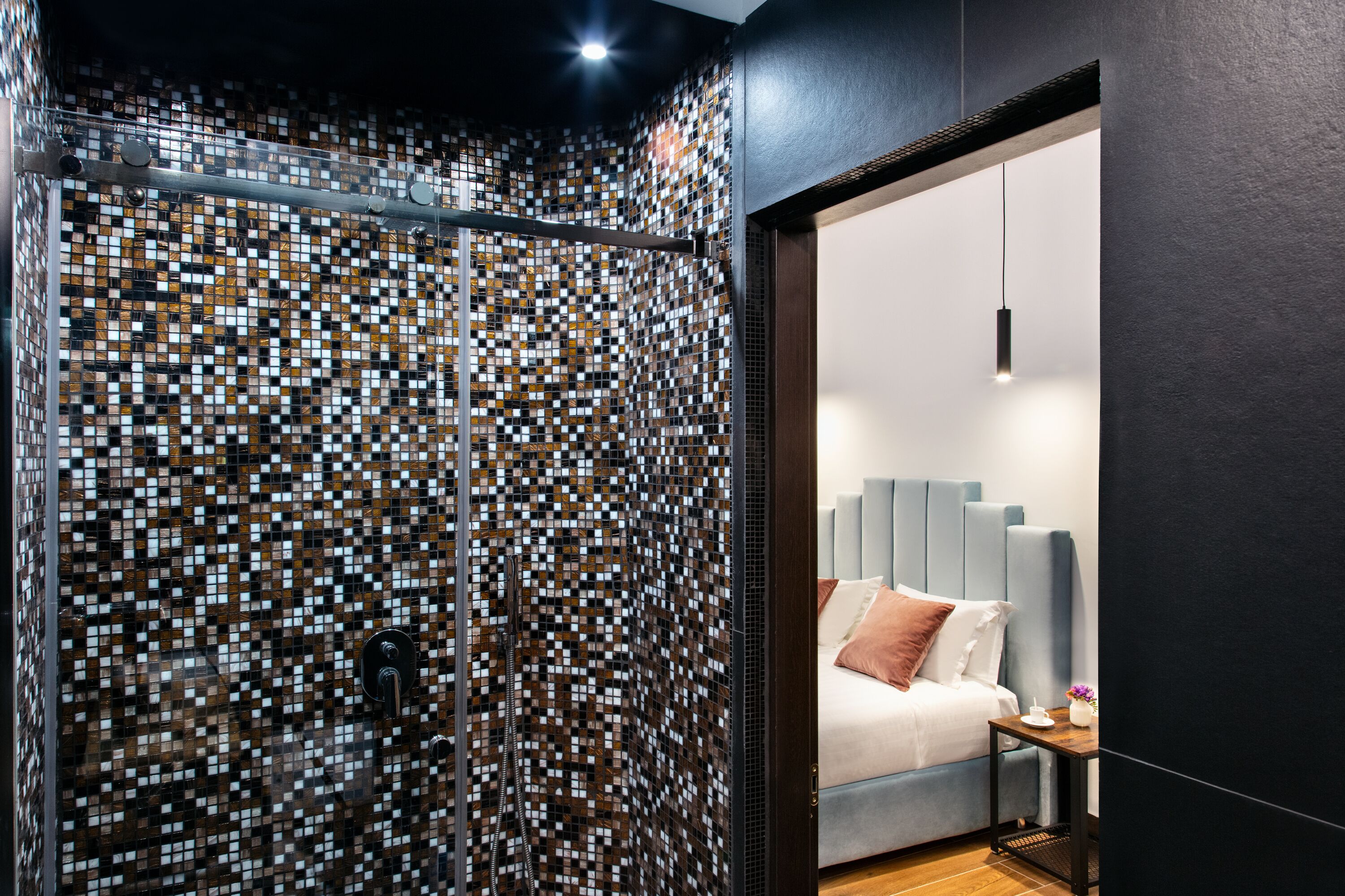 A modern bathroom with a mosaic tile wall, a shower area, and a visible bed with a grey headboard in the adjacent room.