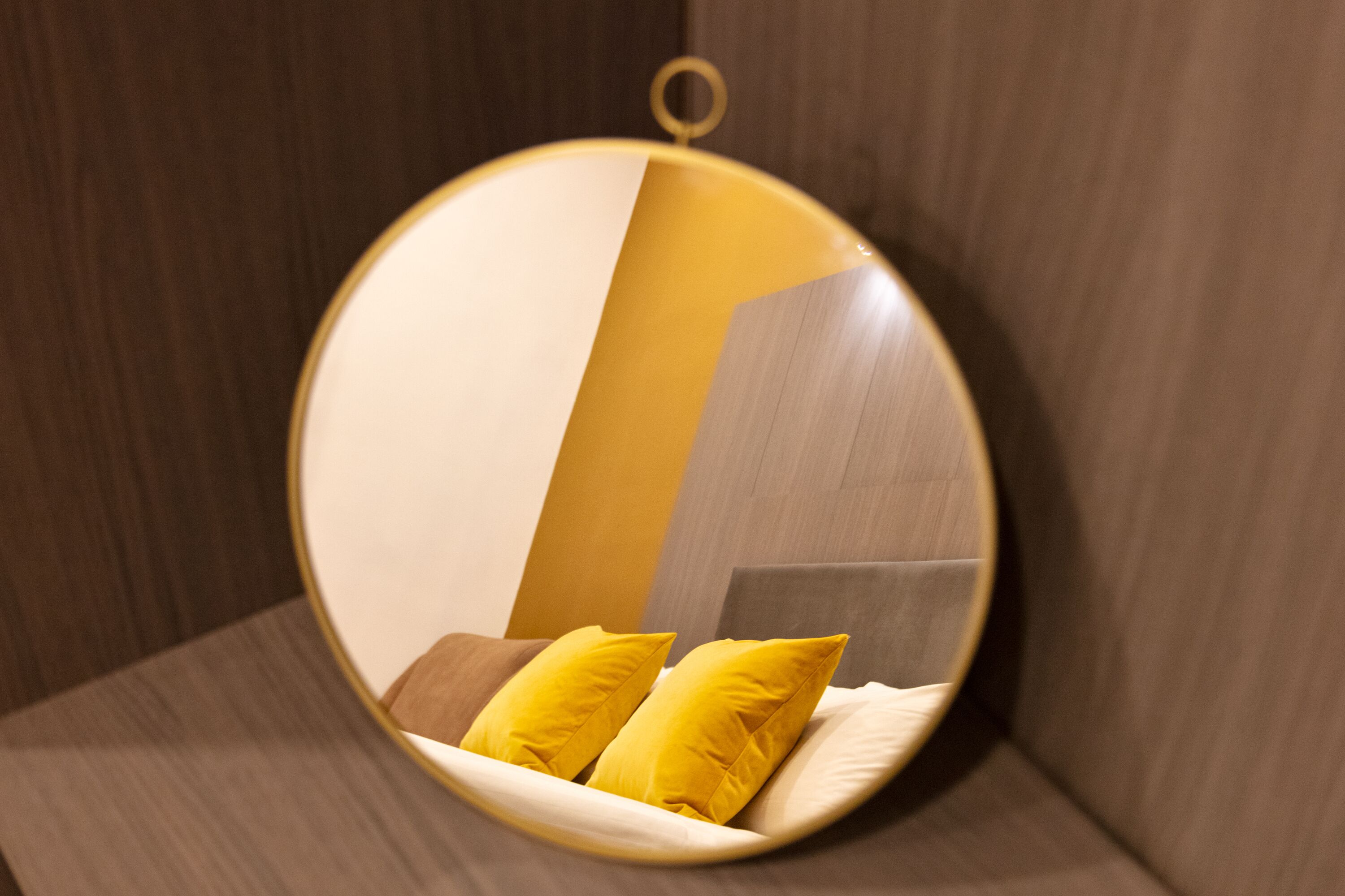 Un miroir rond, doté d’un cadre doré, reflète un lit avec des coussins jaunes et une tête de lit en bois.