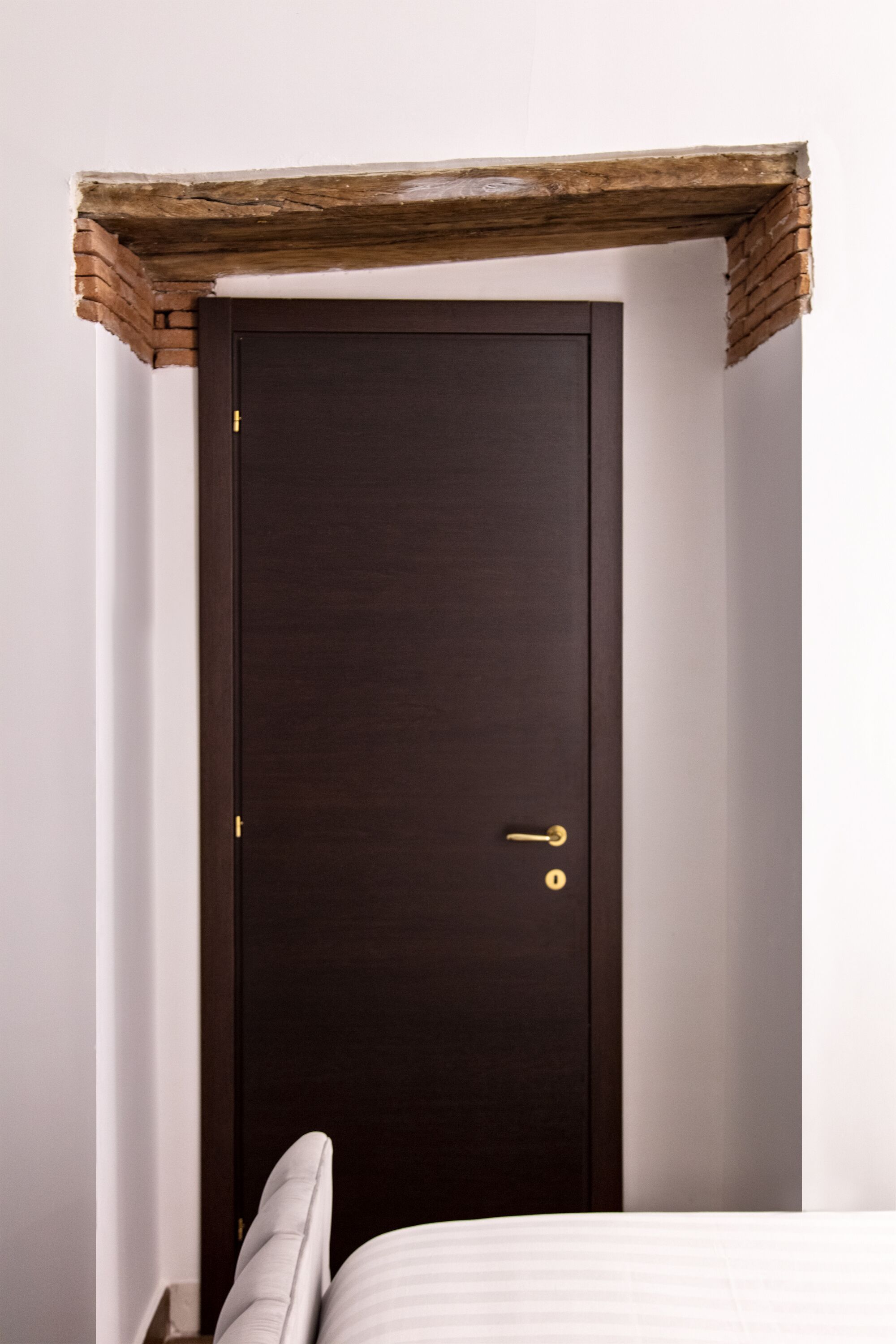 Una puerta de madera oscura con un picaporte dorado y una cerradura pequeña, en una pared blanca con una viga de madera encima.