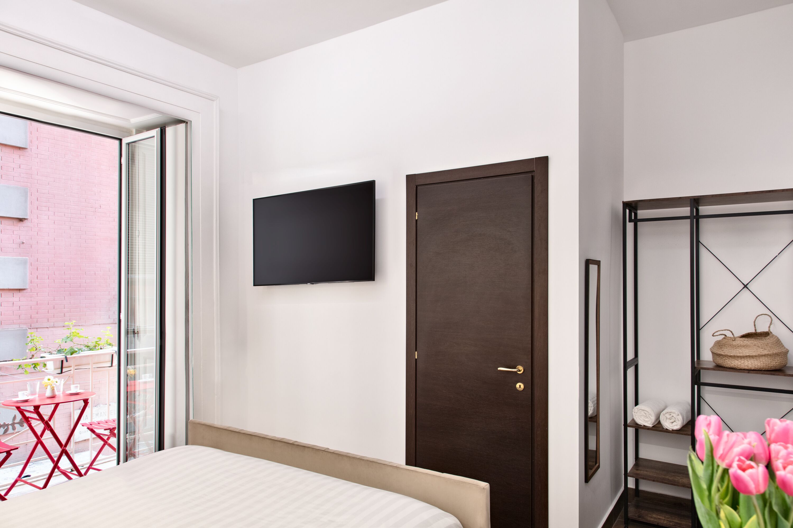 Un dormitorio moderno con una cama, un televisor montado en la pared, una puerta y un estante con toallas y una cesta.