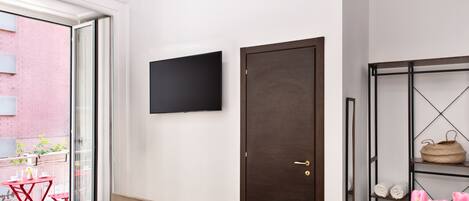 Un dormitorio moderno con una cama, un televisor montado en la pared, una puerta y un estante con toallas y una cesta.