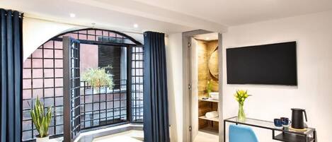 A hotel room with a bed, a desk with a chair, a TV mounted on the wall, and a view through a window with curtains.