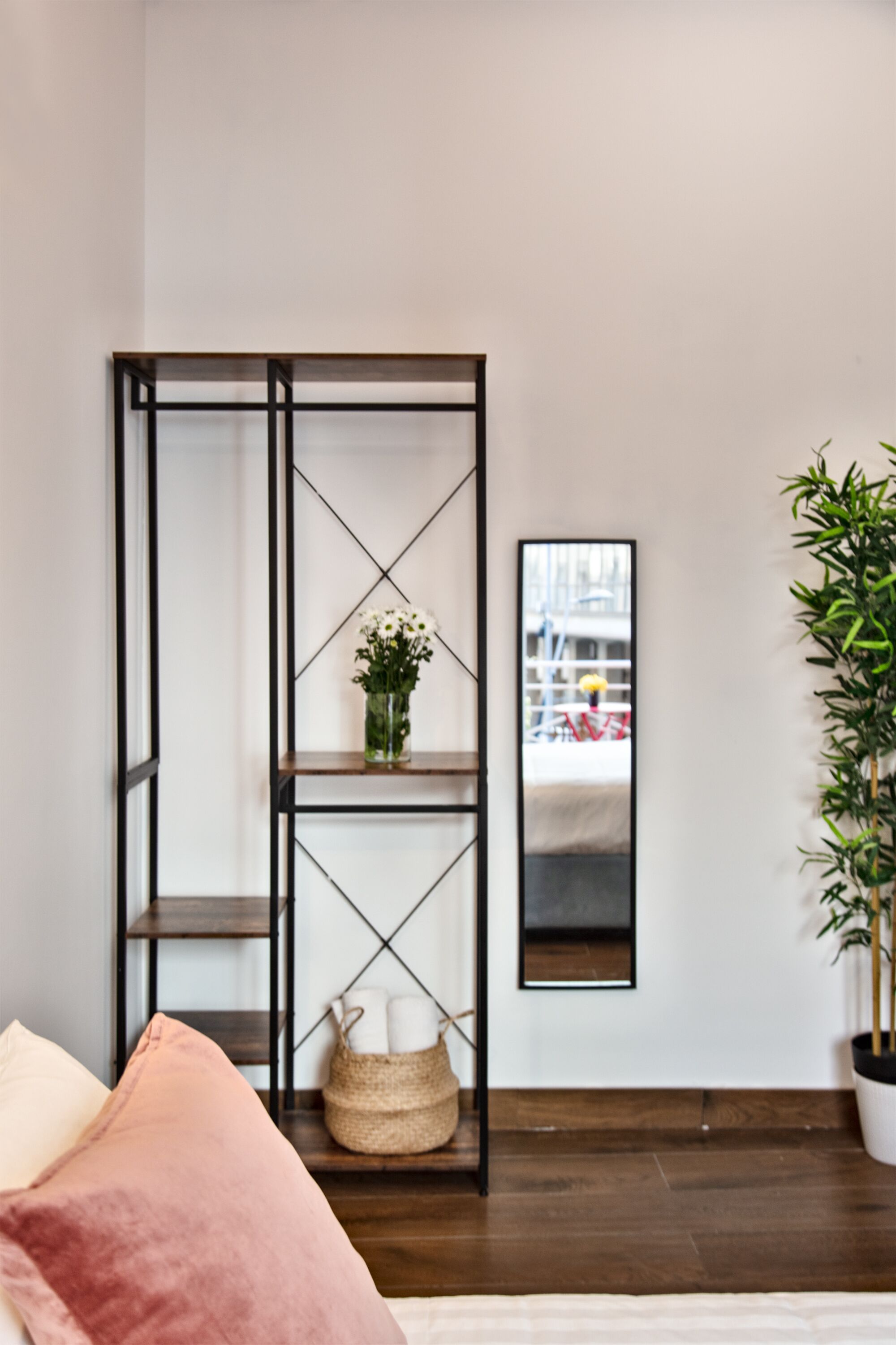 A room with a wooden floor, a bed with pink bedding, a black metal shelf unit with a vase of flowers, a mirror on the wall, and a potted plant.