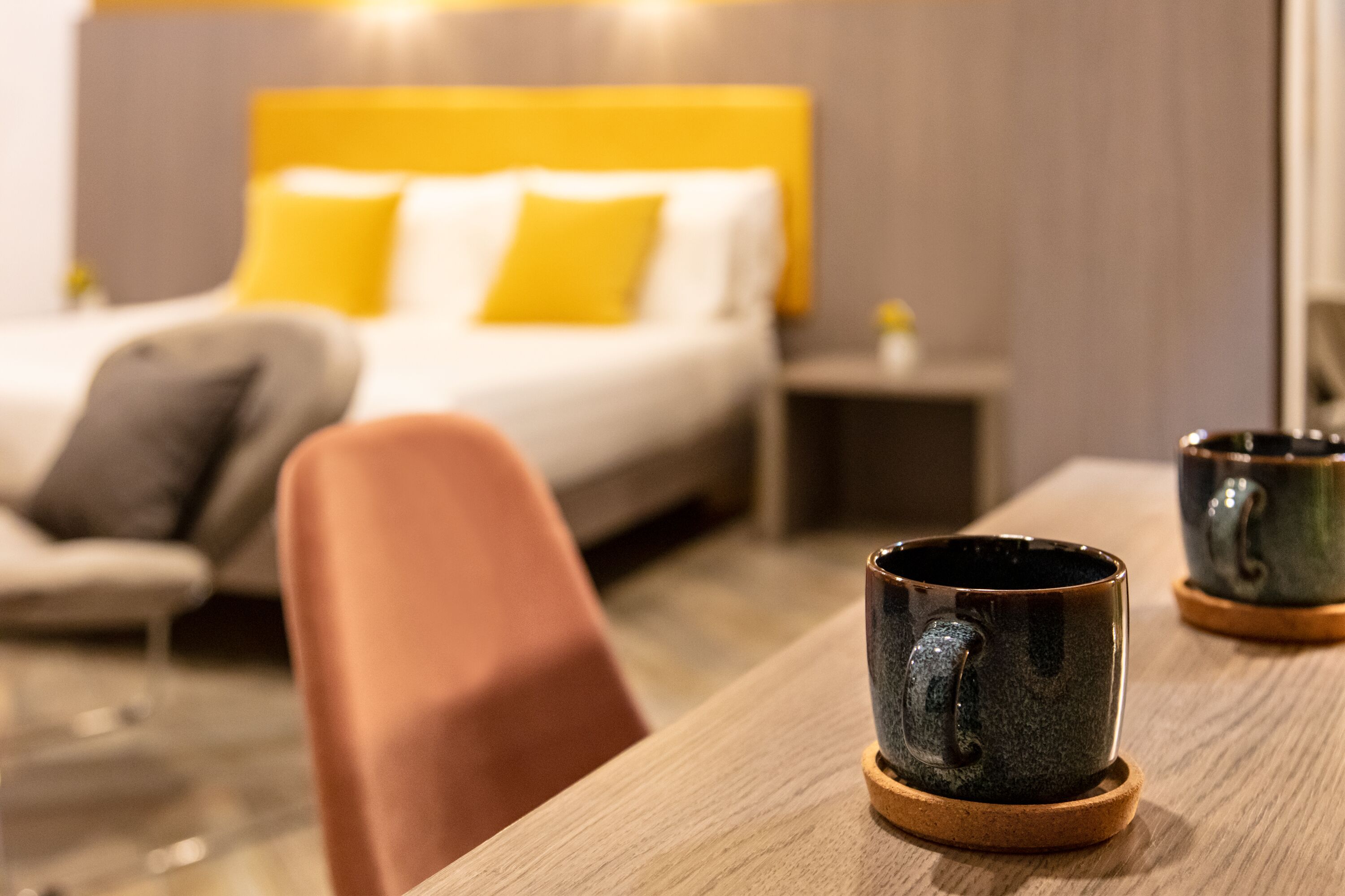 A hotel room with a bed, a nightstand, and two ceramic mugs on a wooden table.