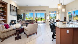 A spacious living area with a fireplace, comfortable seating, a kitchen island, and large windows offering a view of the outdoors. - Private Pool Home in Highly Sought After Mountain View Country Club (La Quinta)