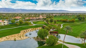 A golf course with a pond, palm trees, and a building in the background. - Private Pool Home in Highly Sought After Mountain View Country Club (La Quinta)