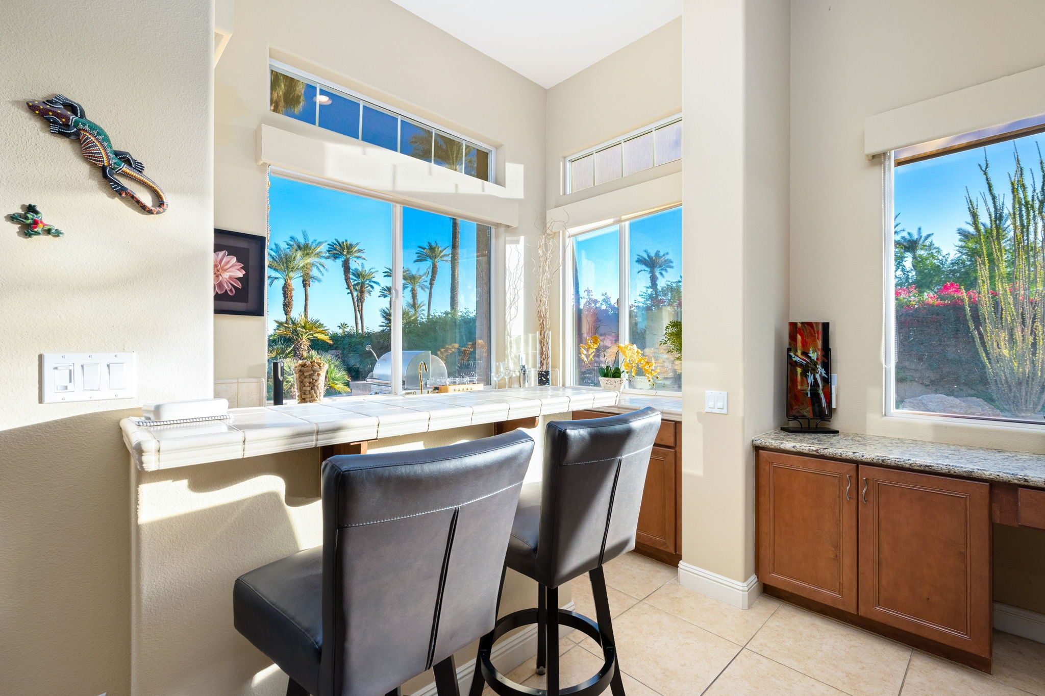 A kitchen with a bar area, large windows with a view of palm trees, and a dining area with two chairs.