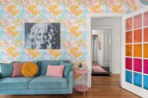 Ein blaugrünes Sofa mit drei Kissen, ein kleiner rosafarbener Beistelltisch und ein farbenfrohes abstraktes Wandbild.