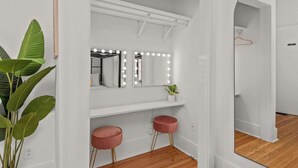 A room with a vanity area featuring a mirror with lights, a shelf, and two stools.