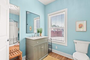Ein Badezimmer mit hellblauer Wand, einer weißen Toilette, einer Holzbank, einem Waschbecken mit Spiegel und einem Fenster mit Jalousien.