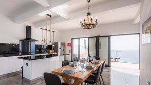 Une cuisine moderne dotée d’un îlot central, d’un coin repas avec une table en bois et d’une vue sur la mer grâce à de grandes fenêtres.