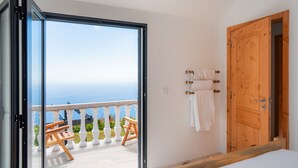 Une pièce dotée d’une grande porte-fenêtre coulissante donnant sur un balcon avec vue sur la mer. Une porte en bois se trouve à droite, et il y a un lit avec une tête de lit en bois.