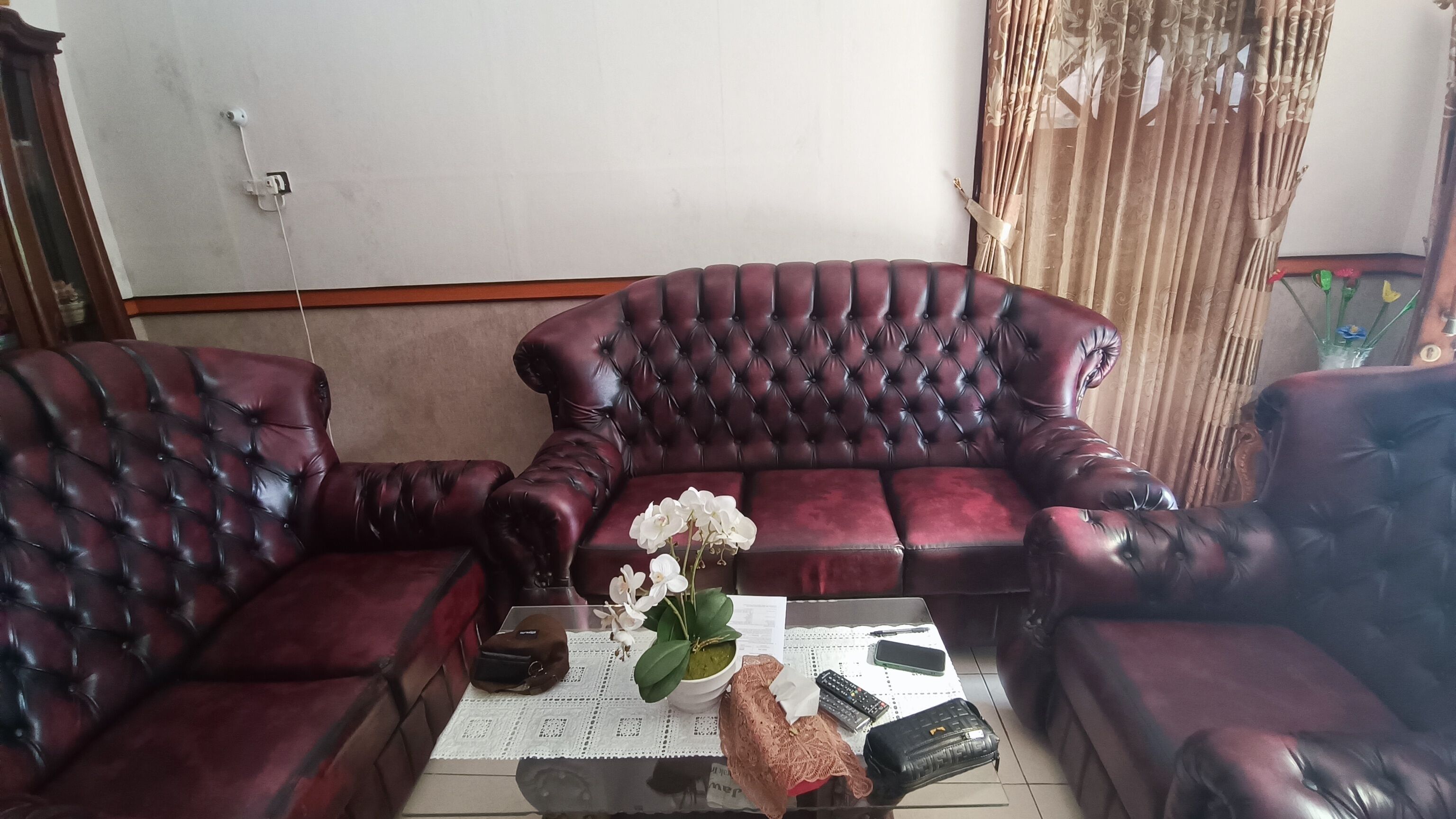 A living room with a dark red leather sofa, a glass coffee table with a lace tablecloth, and a vase with white flowers.