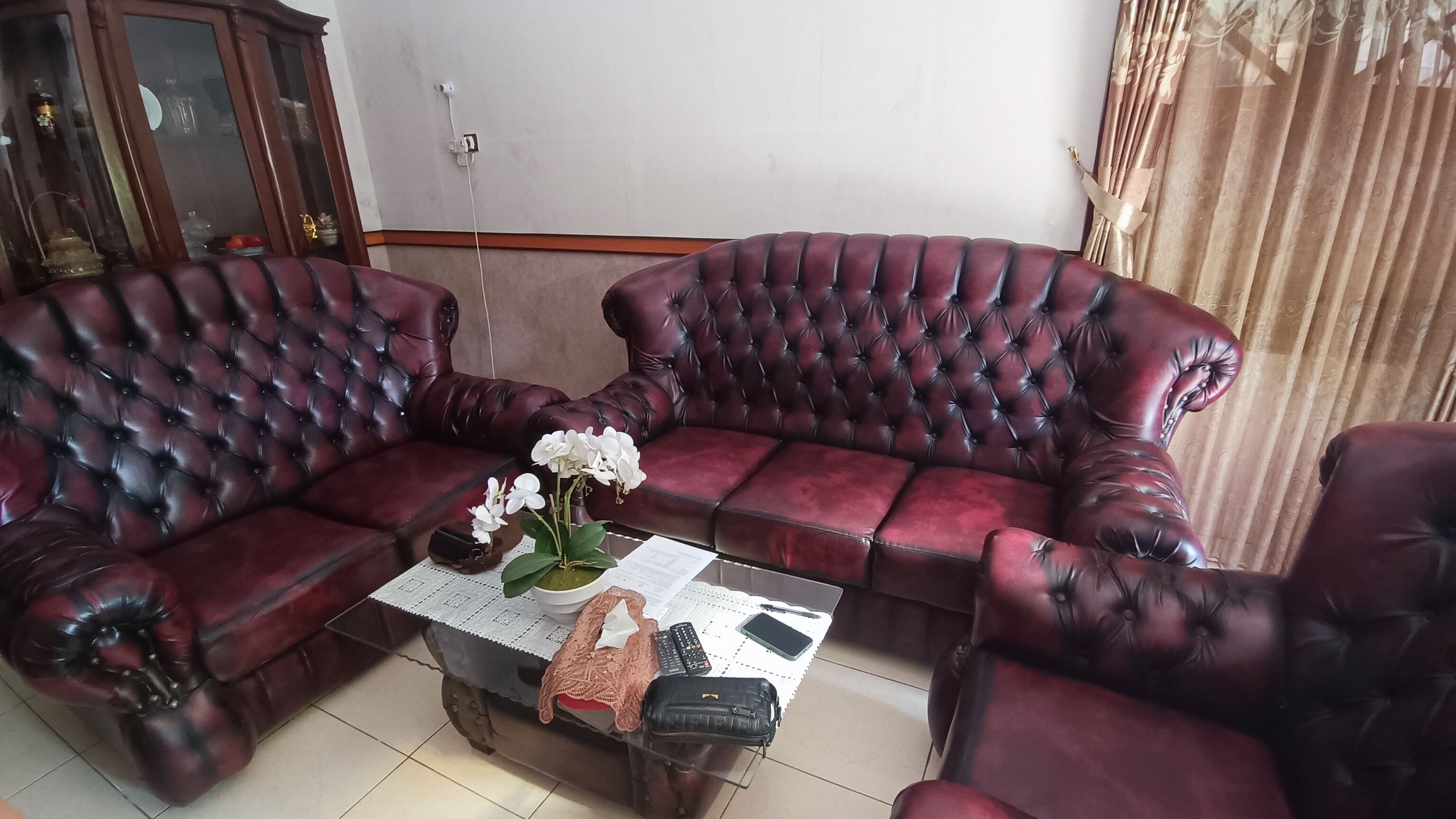 A living room with two dark red leather sofas, a coffee table with a vase of white flowers, and a small table with a remote control, a smartphone, and a notebook.