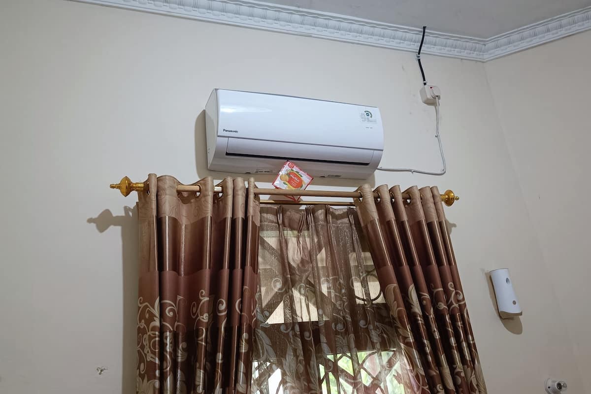 A room with a ceiling-mounted air conditioner, a window with decorative curtains, and a wall-mounted light switch.