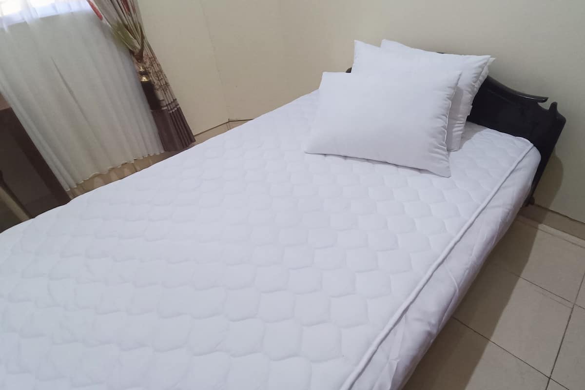 A neatly made bed with a white quilted bedspread and two pillows, a dark headboard, and a plain wall with a window and curtains.