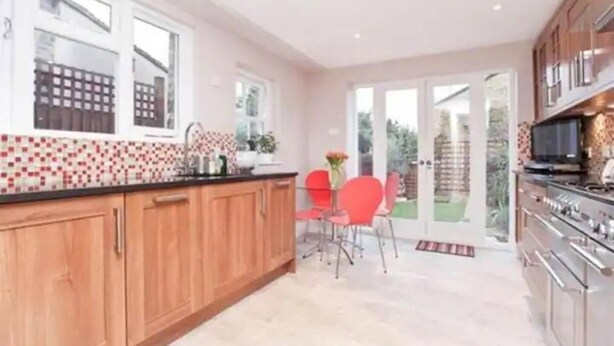 A kitchen with wooden cabinets, a tiled backsplash, a dining area with red chairs, and a sliding glass door leading to a garden. - Luxurious Double Studio in London (London)
