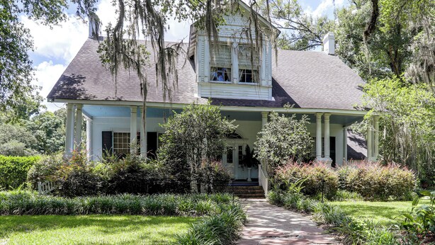 A two-story house with a porch, surrounded by lush greenery and trees.
