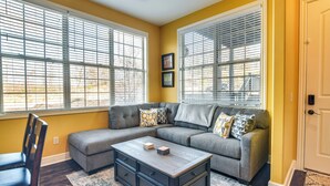 A living room with a sectional sofa, a coffee table, and a chair. There are two windows with blinds, a door, and a small rug on the floor.