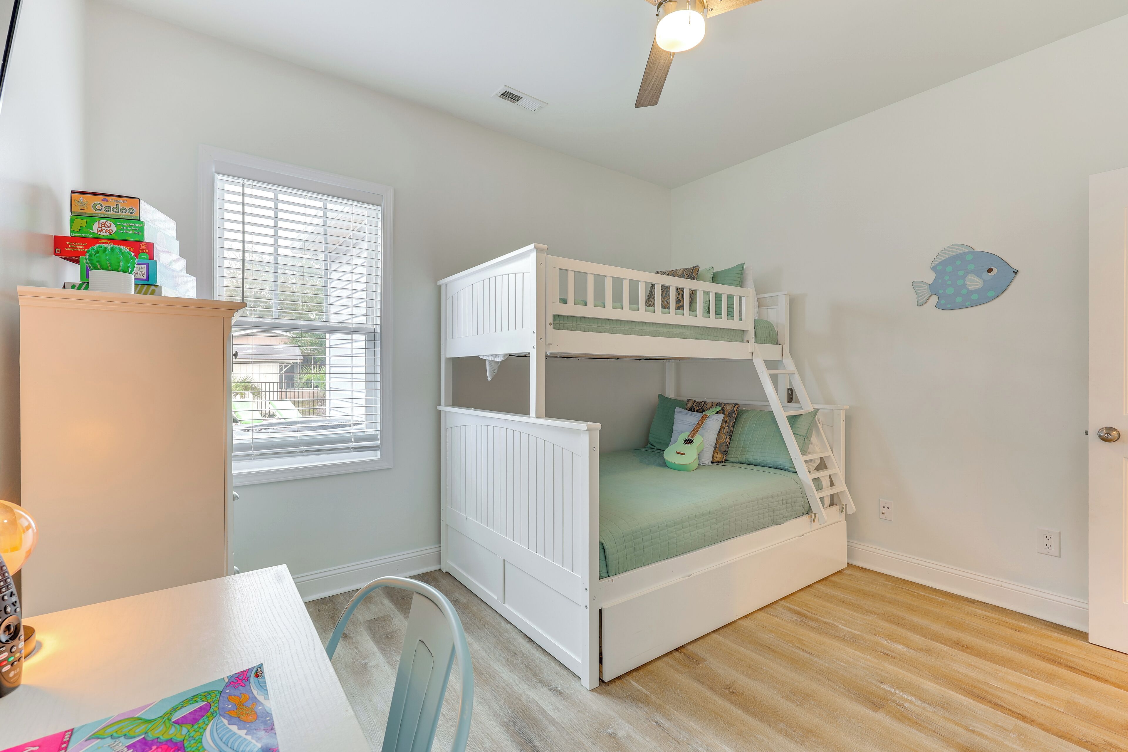 A child's bunk bed with a ladder, a desk with a chair, a window with blinds, and a ceiling fan.