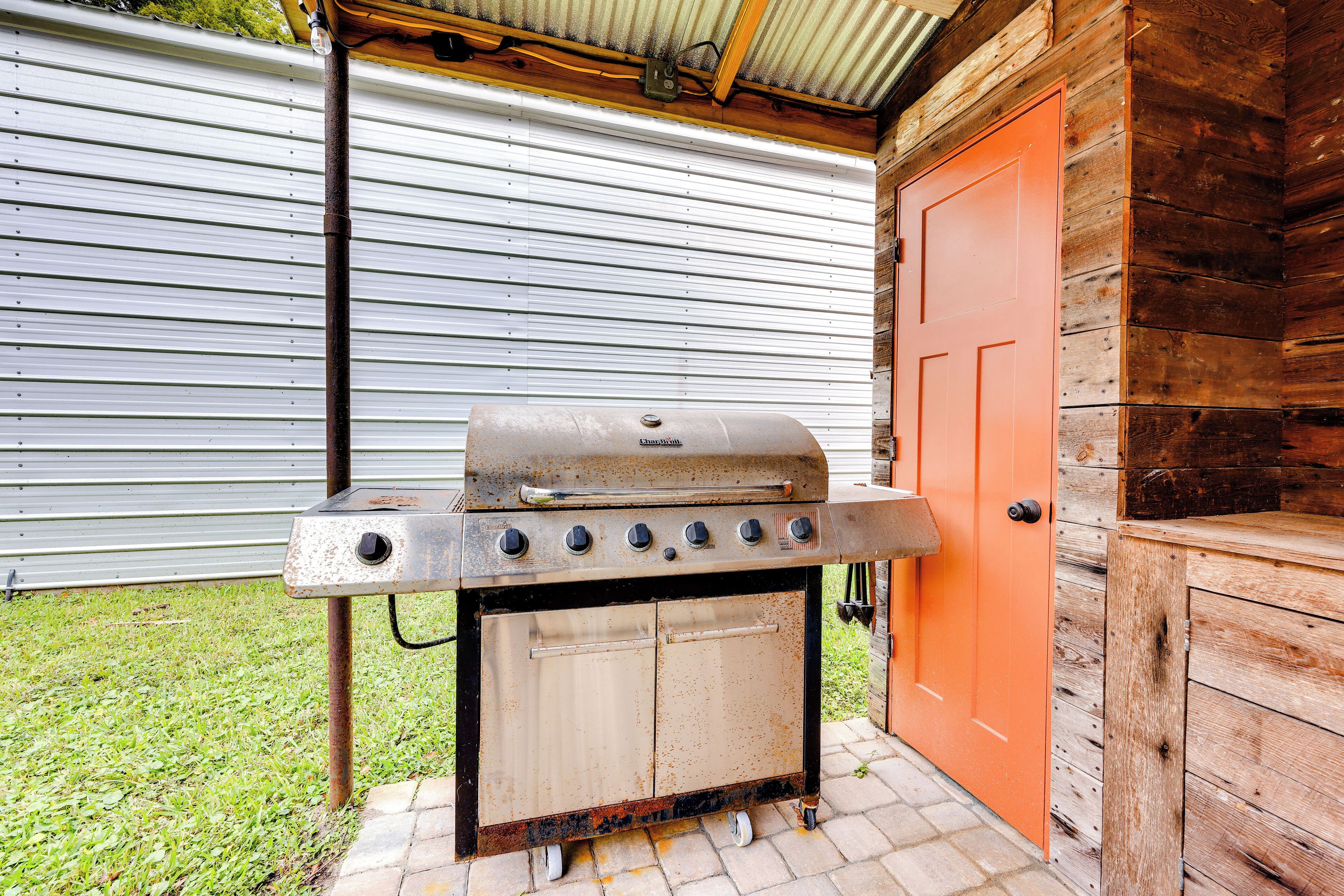 A barbecue grill with a metal hood and four burners, positioned on a brick patio with a wooden door and metal siding in the background.