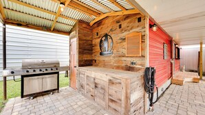Un patio exterior con una estación de parilla de madera, un edificio rojo al fondo y un área cubierta con techo de lámina corrugada.
