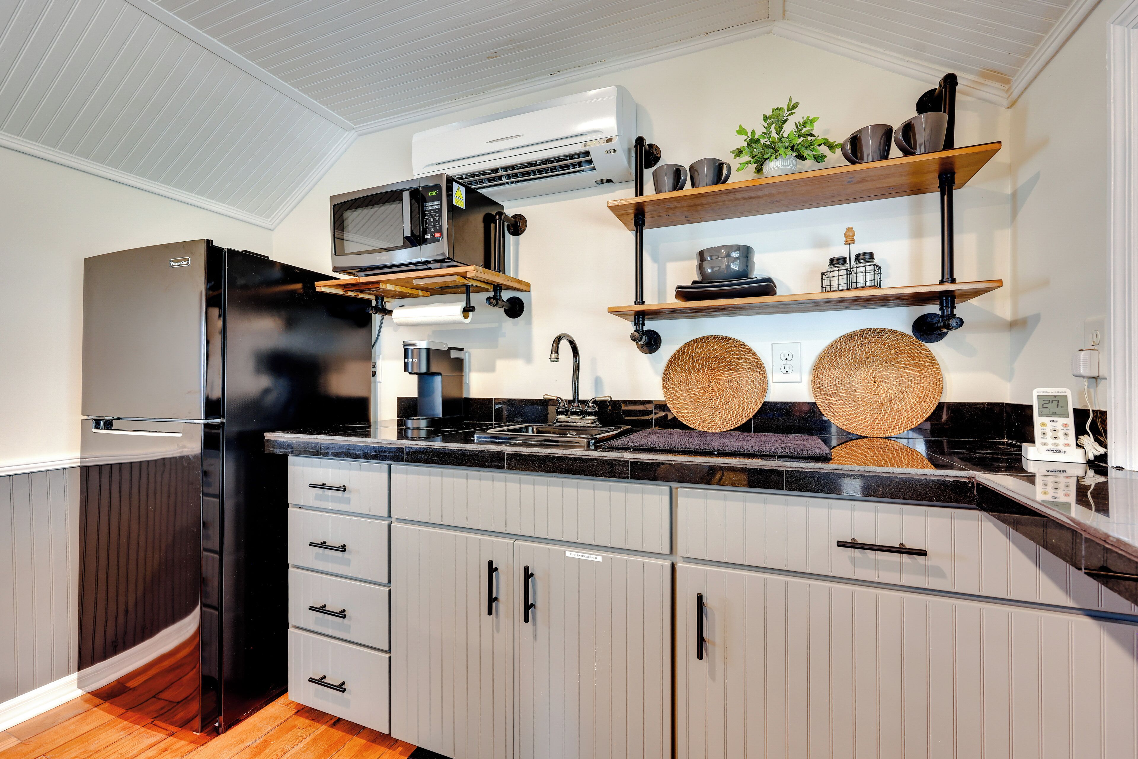 Una cocina moderna con un refrigerador de acero inoxidable, un microondas y un horno empotrado. Hay estantes de madera sobre la encimera con utensilios de cocina y una planta en maceta.