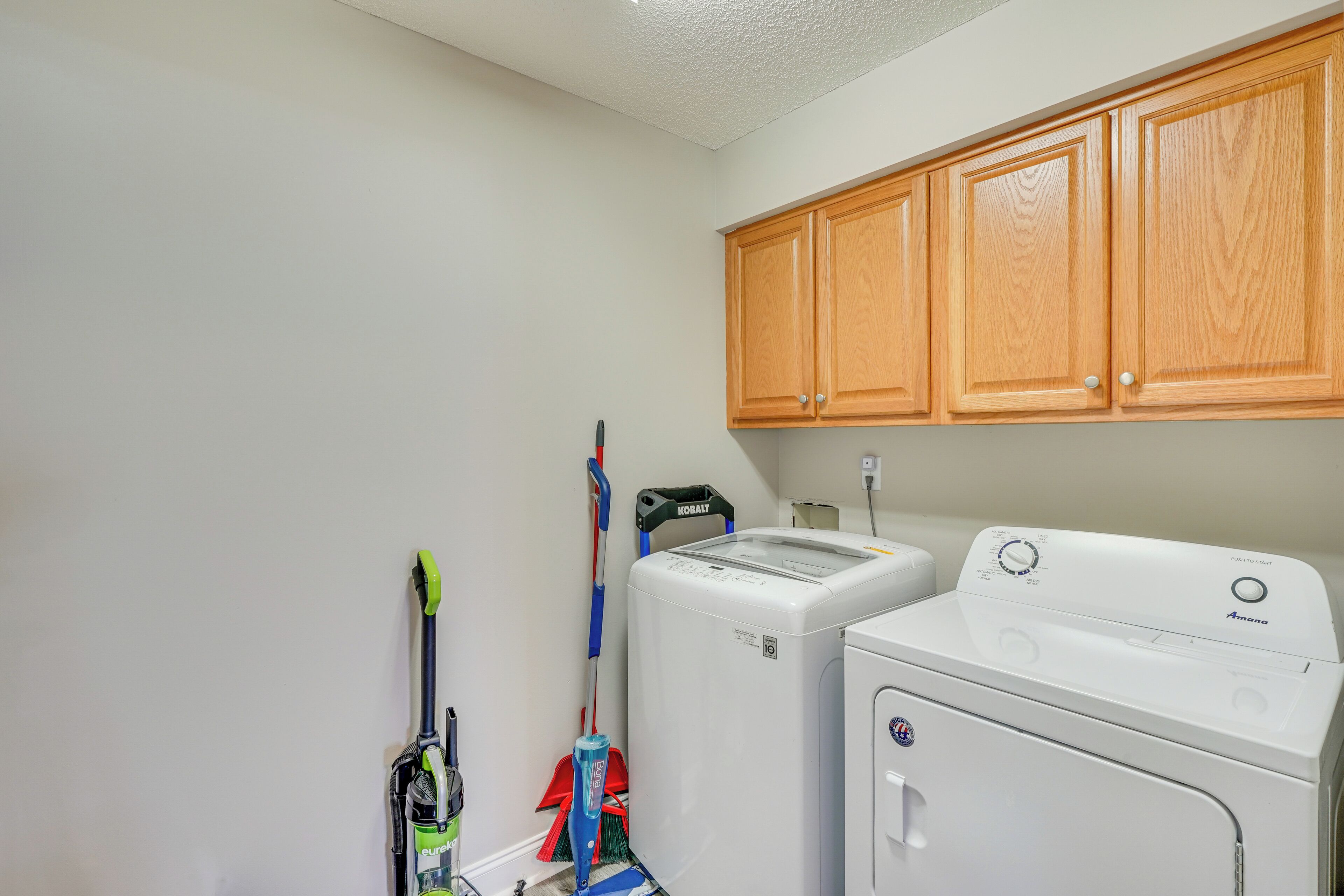 A laundry area with a washing machine and dryer, wooden cabinets above, and cleaning tools hanging on the wall.