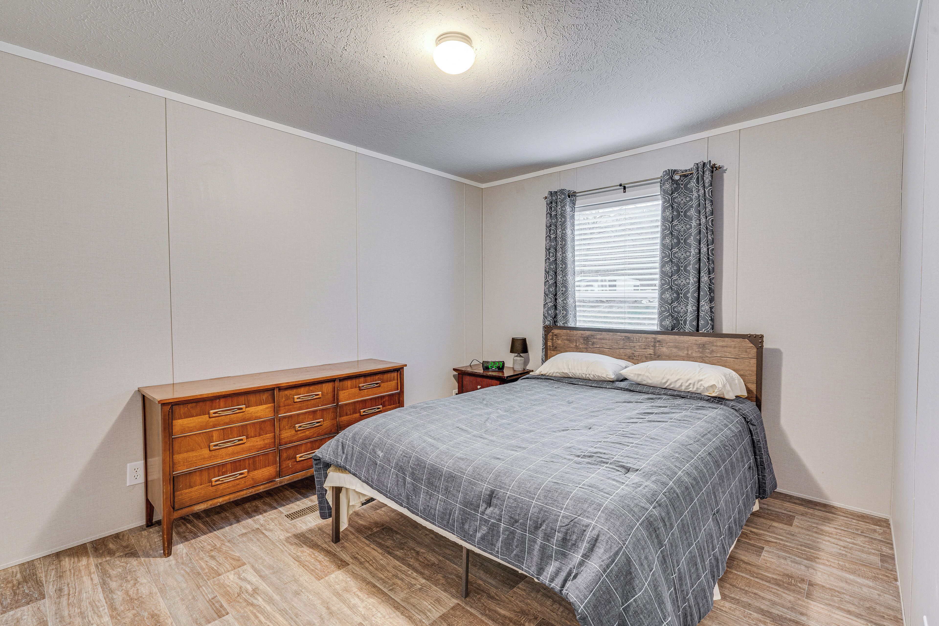 A bedroom with a wooden dresser, a bed with a grey bedspread, a nightstand with a lamp, and a window with patterned curtains.
