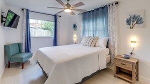 Ein Schlafzimmer mit Bett, Nachttisch, Deckenventilator, an der Wand montiertem Fernseher und einem Fenster mit Vorhängen.