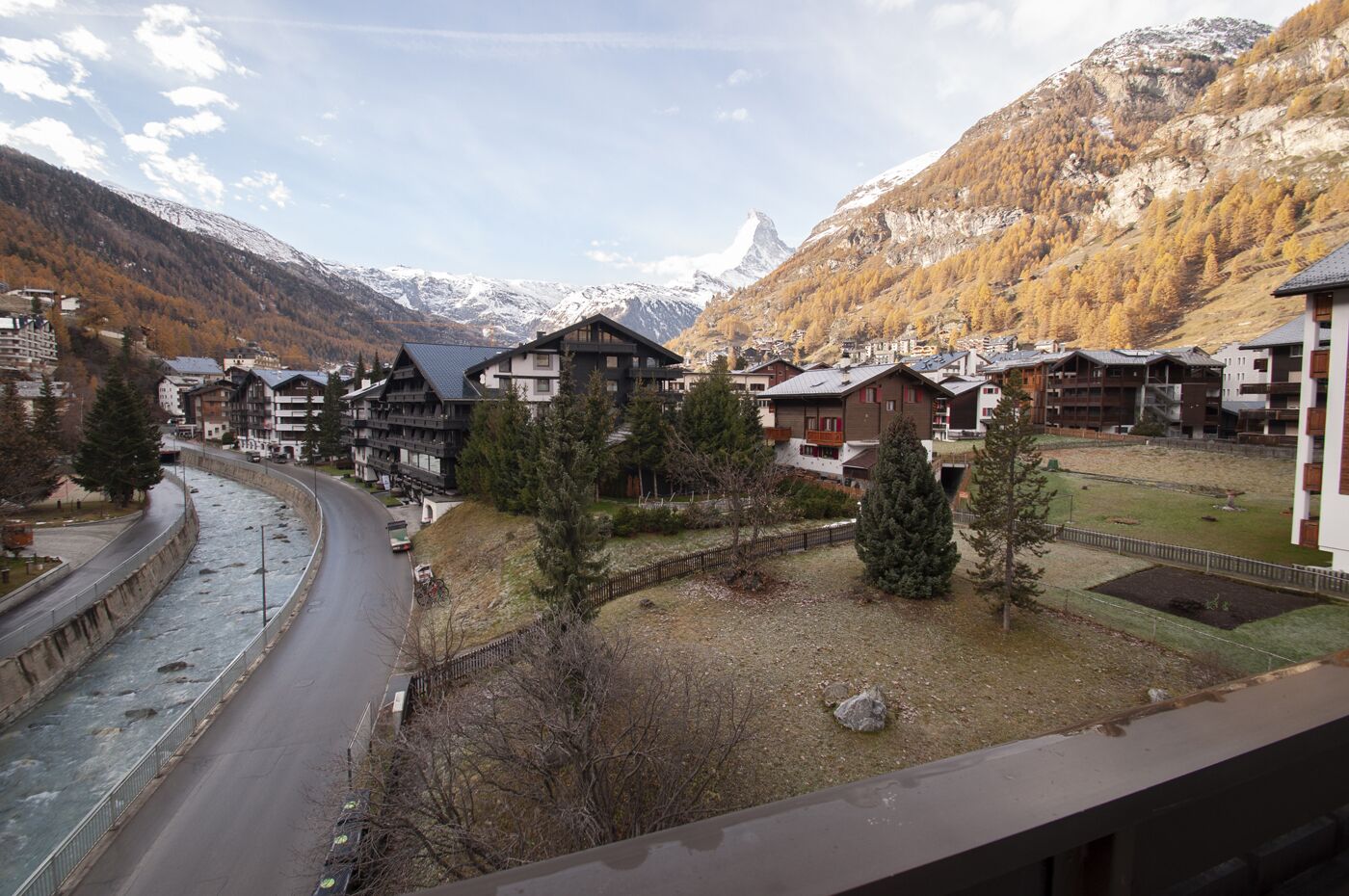 A mountain village with traditional wooden buildings, a river, and snow-capped mountains in the background.