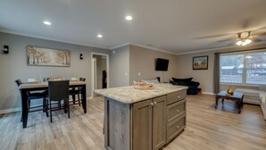 A modern kitchen with a central island, dining area with chairs, a sofa, and a TV mounted on the wall. - Family-friendly Home w/ Deck Near Colonial Beach! (Colonial Beach)